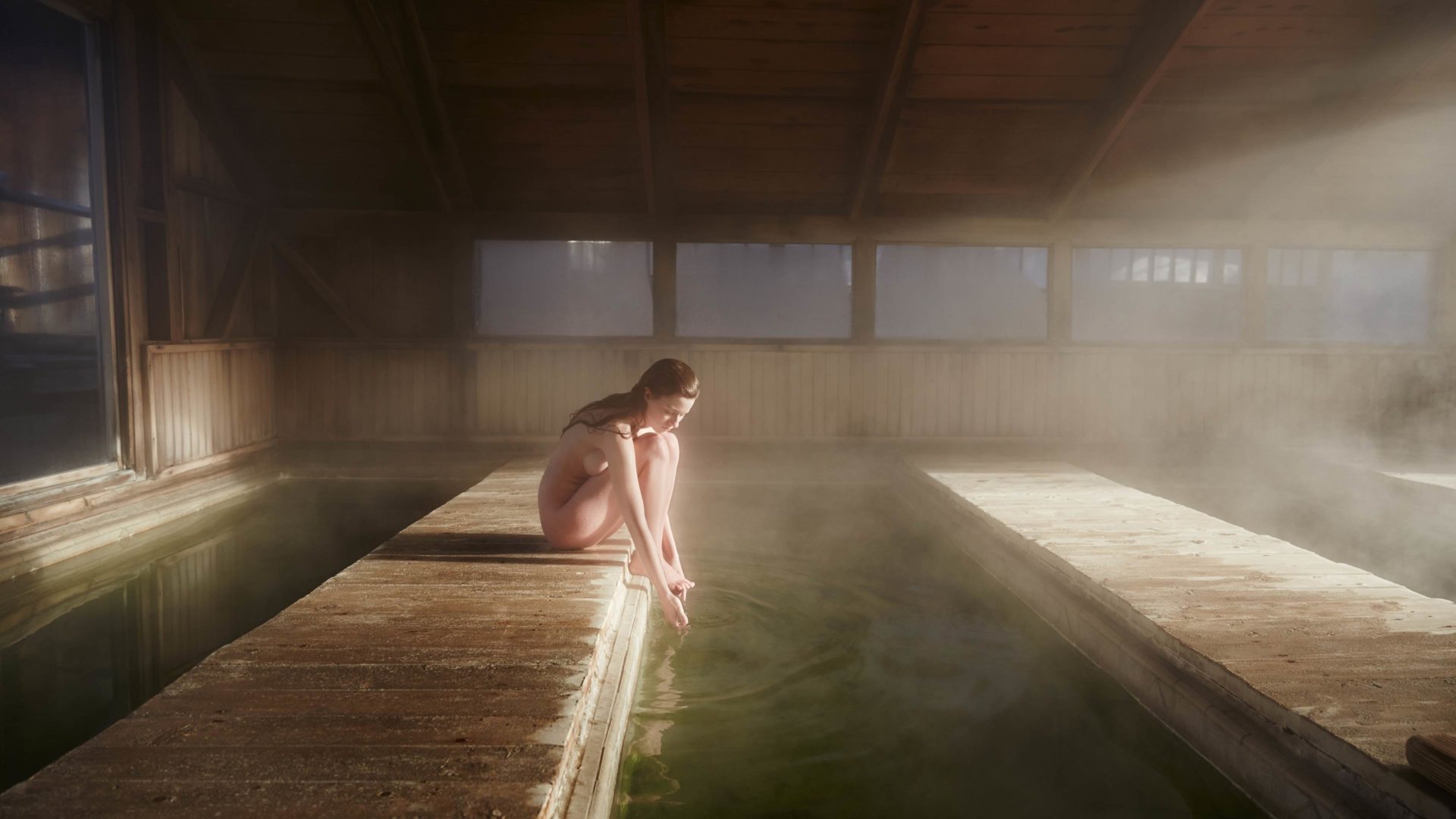 Women sits naked by a green pool, the warm water is steaming