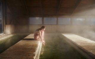Women sits naked by a green pool, the warm water is steaming