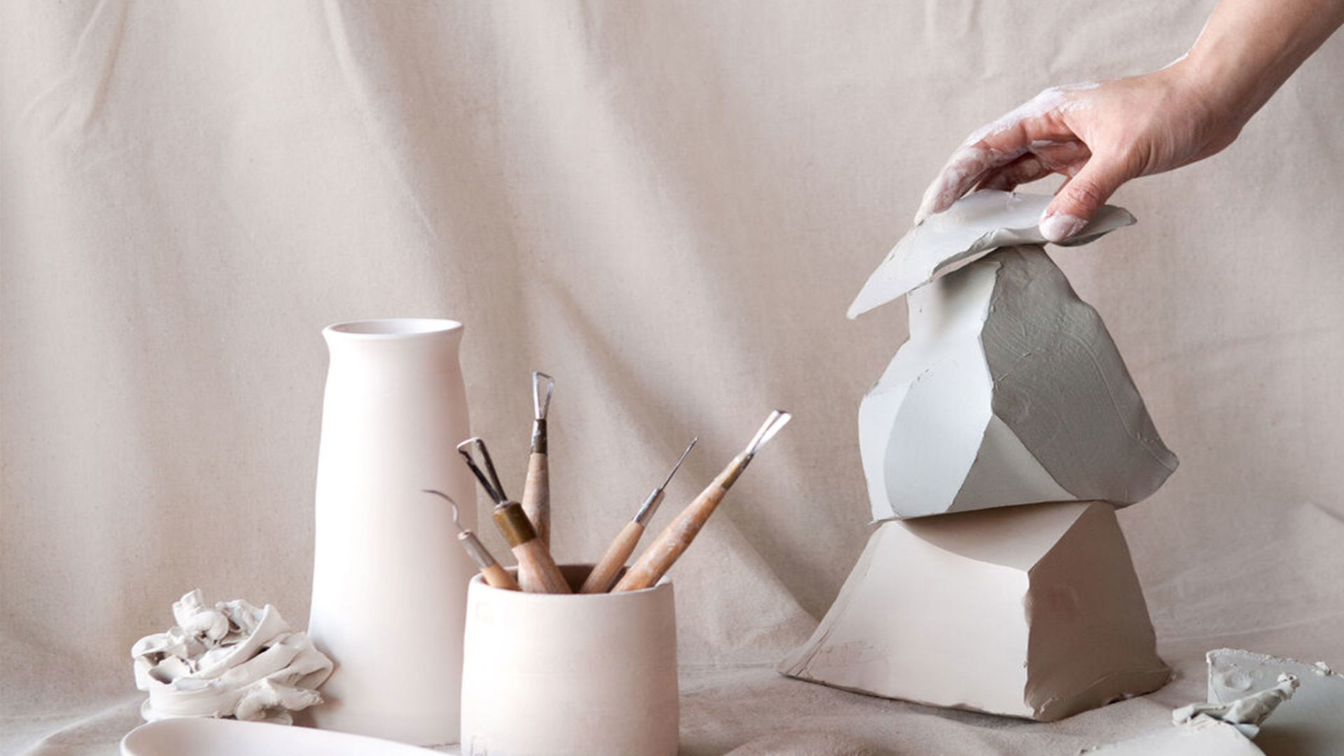 A hand arranges ceramic sculptures alongside artist tools against a pastel pink studio backdrop