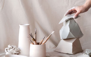 A hand arranges ceramic sculptures alongside artist tools against a pastel pink studio backdrop