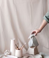 A hand arranges ceramic sculptures alongside artist tools against a pastel pink studio backdrop