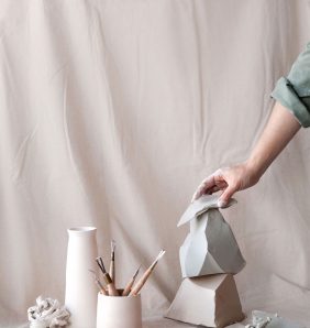 A hand arranges ceramic sculptures alongside artist tools against a pastel pink studio backdrop