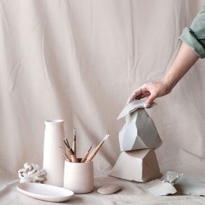 A hand arranges ceramic sculptures alongside artist tools against a pastel pink studio backdrop