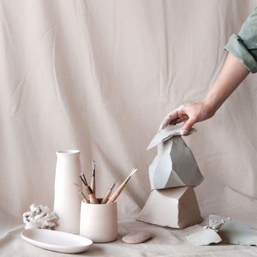 A hand arranges ceramic sculptures alongside artist tools against a pastel pink studio backdrop