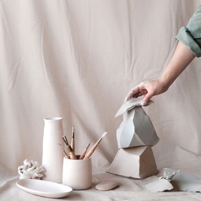 A hand arranges ceramic sculptures alongside artist tools against a pastel pink studio backdrop