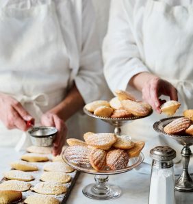 Two chefs prepare and dust dishes of madeleines with sugar