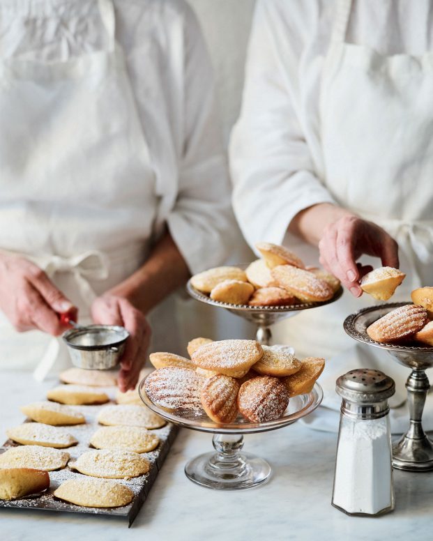 Two chefs prepare and dust dishes of madeleines with sugar