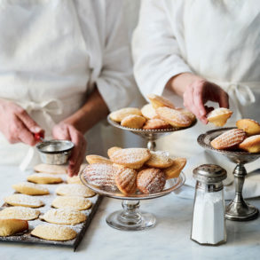 Two chefs prepare and dust dishes of madeleines with sugar